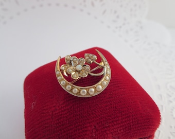 May include: A gold crescent moon brooch with a cluster of white pearls and a single opalescent stone in the center. The brooch is set on a red velvet background.