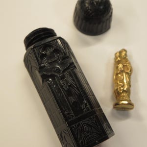 May include: A black perfume bottle with a crucifix relief and gothic details. The bottle is rectangular. A separate black cap and a small gold statue are also present.