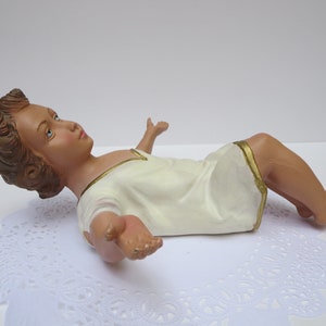 May include: A white baby Jesus figurine with brown hair and blue eyes, lying on its back with arms outstretched. The figurine is wearing a white robe with a gold trim.