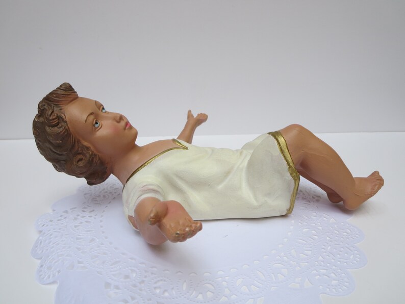 May include: A white baby Jesus figurine with brown hair and blue eyes, lying on its back with arms outstretched. The figurine is wearing a white robe with a gold trim.