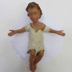 May include: A ceramic figurine of a baby Jesus in a white robe with gold trim, lying on a white lace doily.