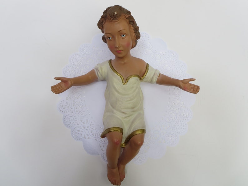 May include: A ceramic figurine of a baby Jesus in a white robe with gold trim, lying on a white lace doily.