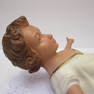May include: A ceramic figurine of a baby Jesus lying down with brown hair, blue eyes, and wearing a white robe with gold trim.