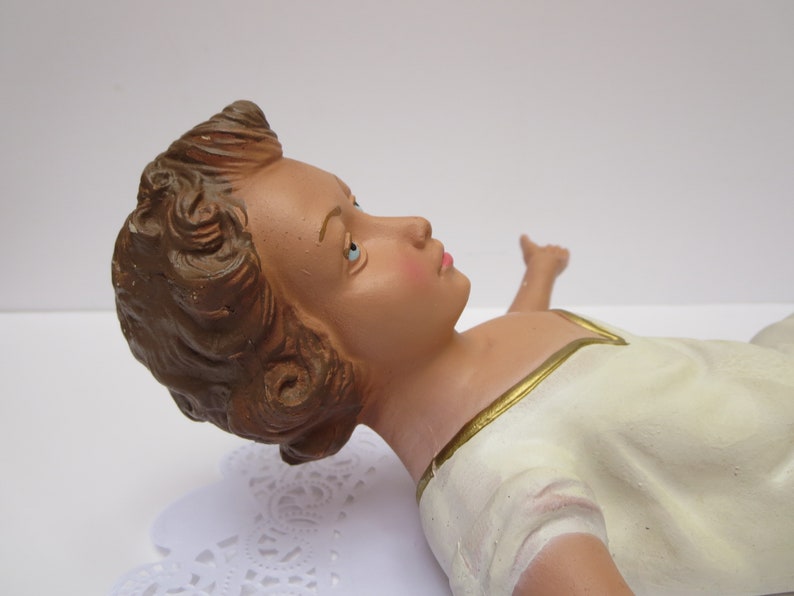 May include: A ceramic figurine of a baby Jesus lying down with brown hair, blue eyes, and wearing a white robe with gold trim.