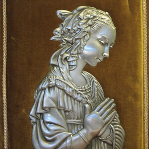 May include: A silver-toned metal wall plaque depicting a woman in a flowing robe with her hands clasped in prayer. The plaque is mounted on a brown velvet background with gold trim.