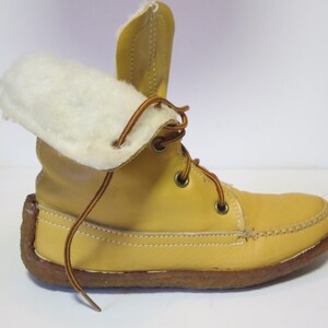 May include: A yellow leather boot with a white, fluffy interior lining. The boot has brown laces and a brown sole. The top of the boot is folded over, revealing the white lining. The boot is designed for cold weather.
