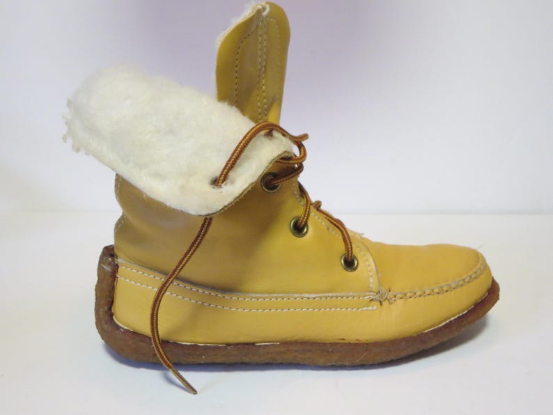 May include: A yellow leather boot with a white, fluffy interior lining. The boot has brown laces and a brown sole. The top of the boot is folded over, revealing the white lining. The boot is designed for cold weather.