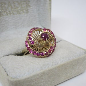 May include: A gold ring featuring a large red ruby surrounded by smaller red rubies. The ring is displayed in a white velvet box.