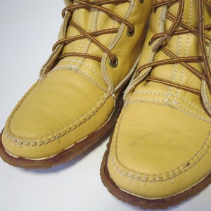 May include: Close-up of two light yellow leather boots with brown laces and stitching. The boots have a moccasin-style toe and a brown rubber sole. The laces are crisscrossed and tied at the top.