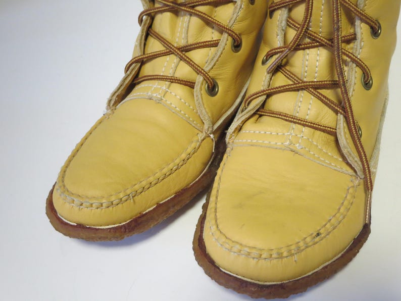 May include: Close-up of two light yellow leather boots with brown laces and stitching. The boots have a moccasin-style toe and a brown rubber sole. The laces are crisscrossed and tied at the top.