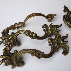 Peut inclure: Ensemble de trois poignées de tiroir en laiton orné avec un motif floral décoratif. Les poignées sont en forme de poignée incurvée avec une extrémité décorative.