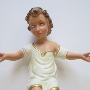 May include: A white plaster figurine of a child with brown hair and outstretched arms. The figurine is wearing a white robe with a gold trim.