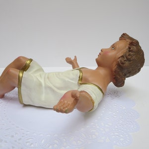 May include: A white baby Jesus figurine with gold trim, lying on its back with its arms outstretched. The figurine is made of plaster and has a realistic appearance.