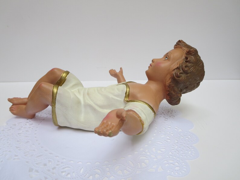 May include: A white baby Jesus figurine with gold trim, lying on its back with its arms outstretched. The figurine is made of plaster and has a realistic appearance.