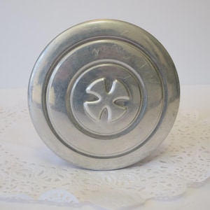 May include: A silver-colored metal lid with a circular design and a cross-shaped emblem in the center. The lid has a slightly worn appearance and sits on a white, lacy doily.