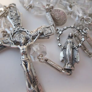 Op de afbeelding: Een zilveren rozenkrans met kristallen kralen. Het middelpunt is een kruisbeeld met een figuur van Jezus Christus en een zilveren hanger van de Maagd Maria.