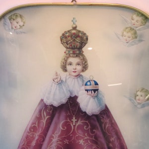 Peut inclure: Une image encadr&eacute;e d'un jeune enfant portant une couronne et une robe rose, tenant un globe. L'enfant est entour&eacute; d'anges avec des ailes.
