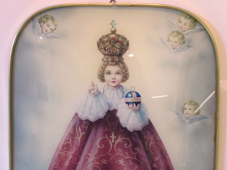 Peut inclure: Une image encadr&eacute;e d'un jeune enfant portant une couronne et une robe rose, tenant un globe. L'enfant est entour&eacute; d'anges avec des ailes.