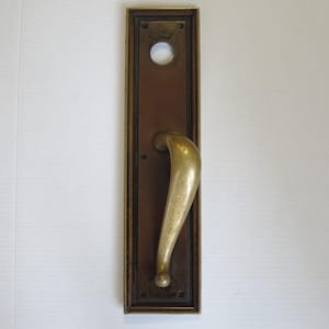 May include: An antique door handle and plate. The rectangular plate is a dark brown with a gold-colored border and a circular hole at the top. The curved handle is gold-colored. The handle is attached to the plate with a small, round knob.