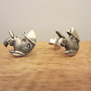May include: A pair of silver earrings featuring a detailed sculpted monkey wearing a hat. The monkey is holding a small object in its hand.