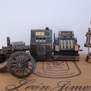 May include: A set of vintage-style pencil sharpeners, including a cannon, a slot machine, a cash register, and a rotary phone. The sharpeners are made of metal and have a bronze finish. The cash register has the text "Cash Sales" and "National" on it.
