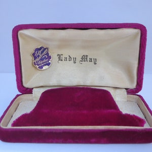 May include: Open magenta velvet jewelry box with a cream-colored satin interior. The box features a magenta velvet insert and the text "Lady May" printed inside. A small pin with the text "Edgar Chalom Bijoutier Montreal" is also visible.