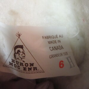May include: A white label with text and a logo, possibly from footwear. The label states "FABRIQU&Eacute; AU MADE IN CANADA GRANDEUR/SIZE 6 H" and features a Native American profile within a triangle.