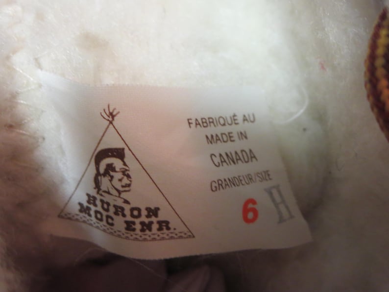 May include: A white label with text and a logo, possibly from footwear. The label states "FABRIQU&Eacute; AU MADE IN CANADA GRANDEUR/SIZE 6 H" and features a Native American profile within a triangle.