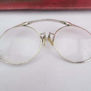 Może przedstawiać: Okulary w srebrnym kolorze z okrągłymi oprawkami i ozdobnym wzorem na ramie. Okulary są połączone zawiasem na środku i mają małą pętelkę po jednej stronie.