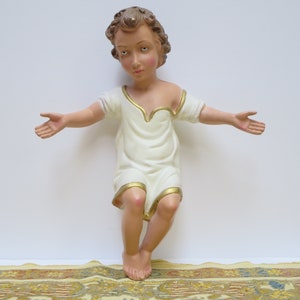 May include: A white figurine of a young child with brown hair and a white robe with gold trim. The child is sitting with arms outstretched, as if in a gesture of blessing.