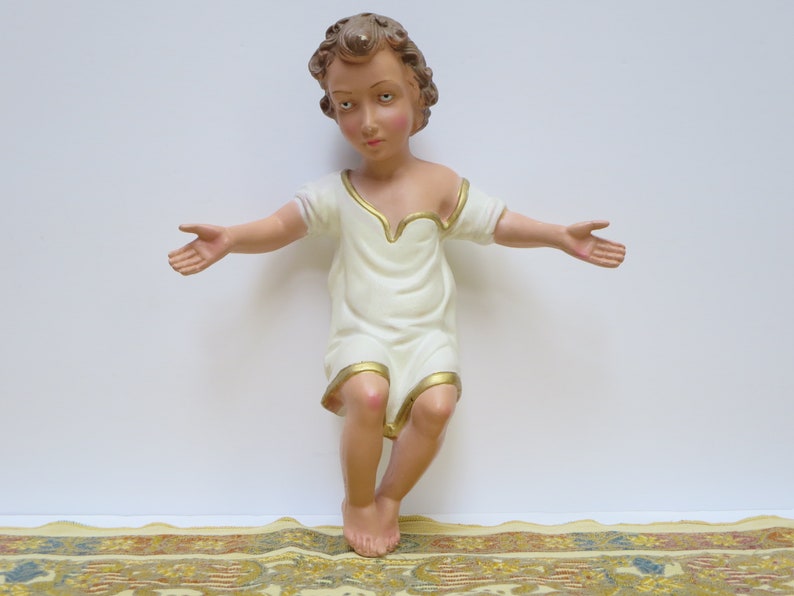 May include: A white figurine of a young child with brown hair and a white robe with gold trim. The child is sitting with arms outstretched, as if in a gesture of blessing.