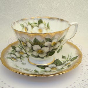Peut inclure: Une tasse à thé et une soucoupe blanches avec un bord doré et un motif floral. Le motif floral présente des fleurs blanches avec des feuilles vertes sur un fond blanc.