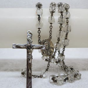 May include: A silver rosary with clear glass beads and a silver crucifix with a figure of Jesus Christ. The crucifix has a small silver medallion with a religious image.