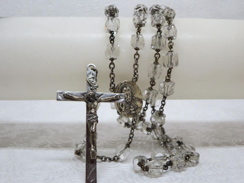 May include: A silver rosary with clear glass beads and a silver crucifix with a figure of Jesus Christ. The crucifix has a small silver medallion with a religious image.