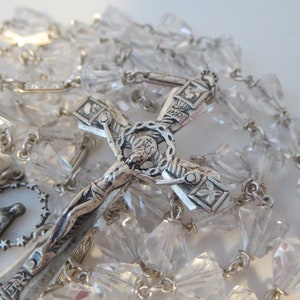 Op de afbeelding: Een zilveren rozenkrans met kristallen kralen. De rozenkrans heeft een kruisbeeld met een figuur van Jezus Christus en een klein zilveren hanger van de Maagd Maria.