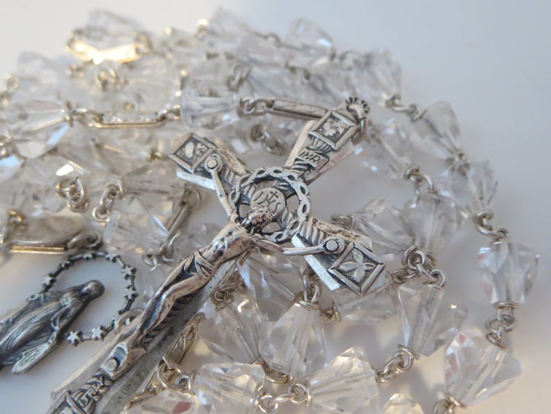 Op de afbeelding: Een zilveren rozenkrans met kristallen kralen. De rozenkrans heeft een kruisbeeld met een figuur van Jezus Christus en een klein zilveren hanger van de Maagd Maria.