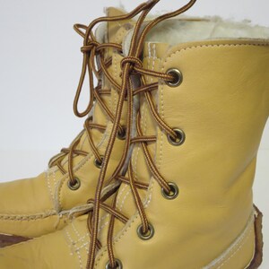 May include: A pair of light tan leather boots with brown laces and metal eyelets. The boots have a soft, white interior lining and a stitched sole. The laces are tied in a crisscross pattern.