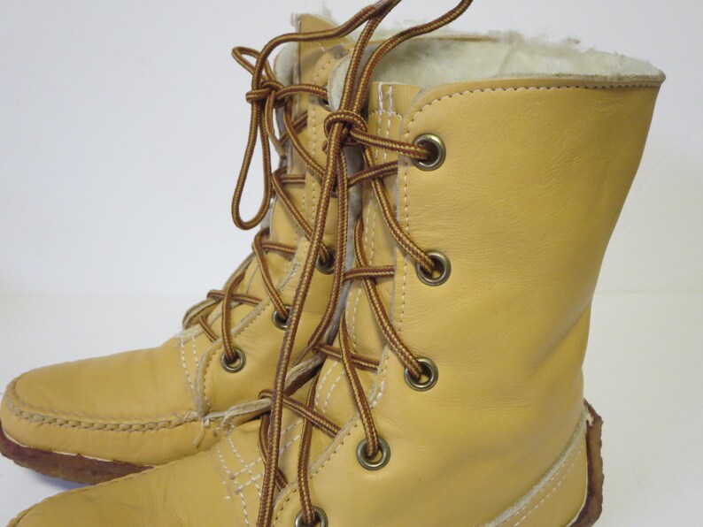 May include: A pair of light tan leather boots with brown laces and metal eyelets. The boots have a soft, white interior lining and a stitched sole. The laces are tied in a crisscross pattern.