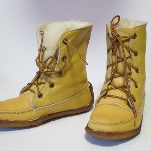 May include: A pair of yellow leather boots with brown laces and a faux fur lining. The boots have a moccasin-style toe and a brown sole. The laces are tied in a crisscross pattern. The boots are mid-calf height.