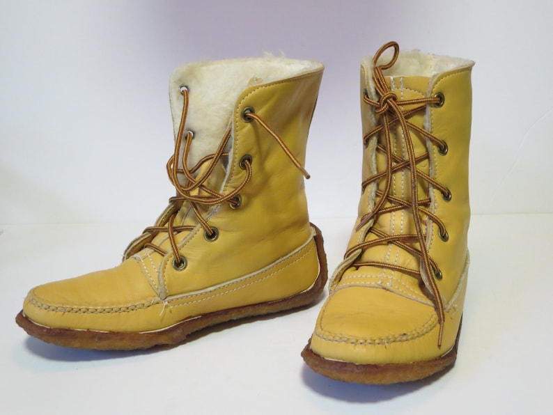 May include: A pair of yellow leather boots with brown laces and a faux fur lining. The boots have a moccasin-style toe and a brown sole. The laces are tied in a crisscross pattern. The boots are mid-calf height.