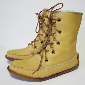 May include: A pair of light yellow leather boots with brown laces. The boots have a moccasin-style toe and a faux fur lining. The soles are brown and the boots are laced up the front. The boots are mid-calf height.