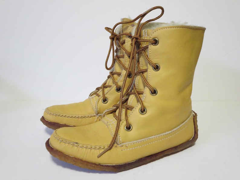 May include: A pair of light yellow leather boots with brown laces. The boots have a moccasin-style toe and a faux fur lining. The soles are brown and the boots are laced up the front. The boots are mid-calf height.