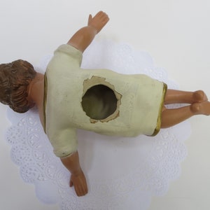 May include: A white plaster figurine of a baby Jesus lying on its back with its arms outstretched. The figurine has a hole in the back, suggesting it may be a nativity scene piece.