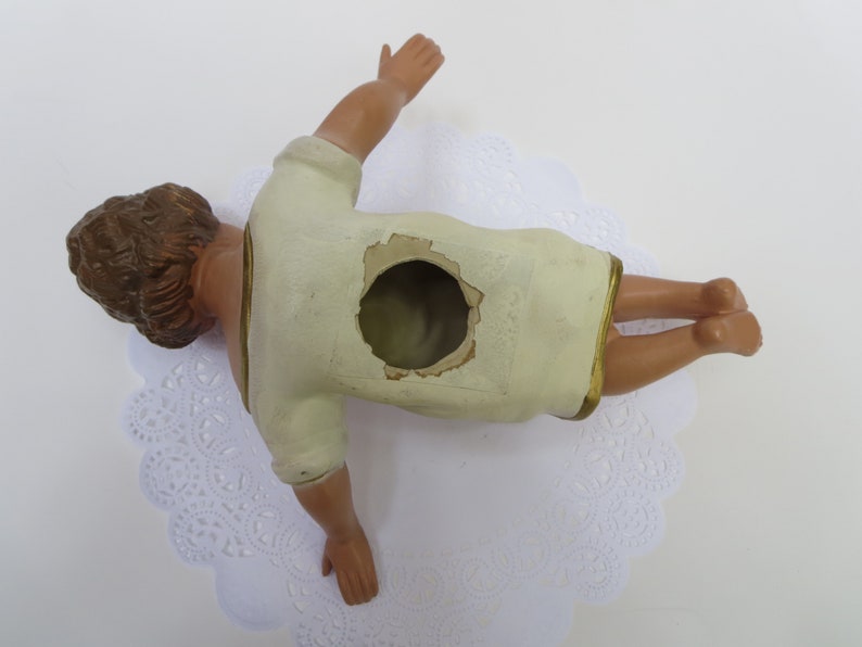 May include: A white plaster figurine of a baby Jesus lying on its back with its arms outstretched. The figurine has a hole in the back, suggesting it may be a nativity scene piece.