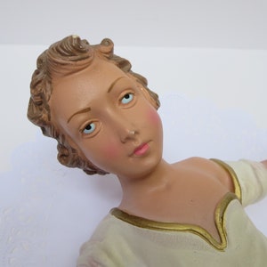 May include: A ceramic figurine of an angel with light brown hair, blue eyes, and a white dress with gold trim.