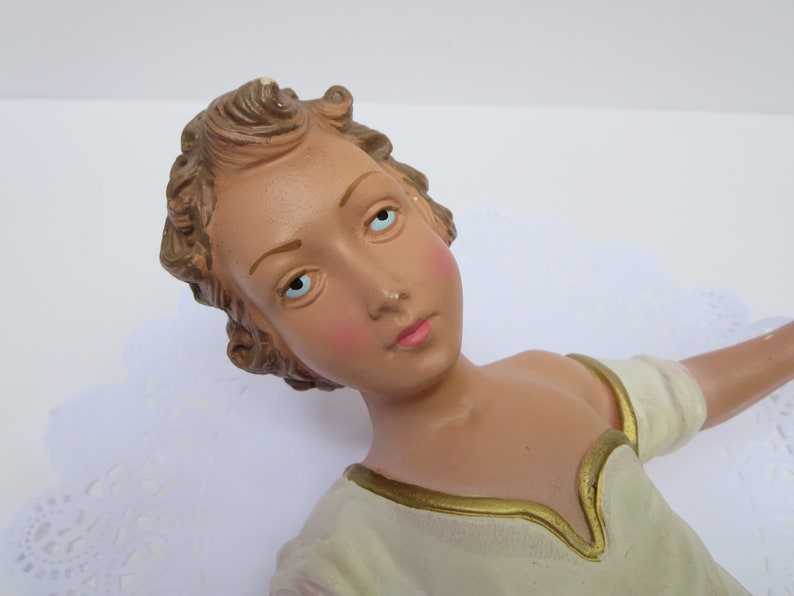 May include: A ceramic figurine of an angel with light brown hair, blue eyes, and a white dress with gold trim.
