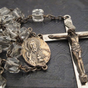 May include: A silver rosary with a clear glass bead chain, a silver crucifix with a figure of Jesus Christ, and a silver medallion with a depiction of a saint.