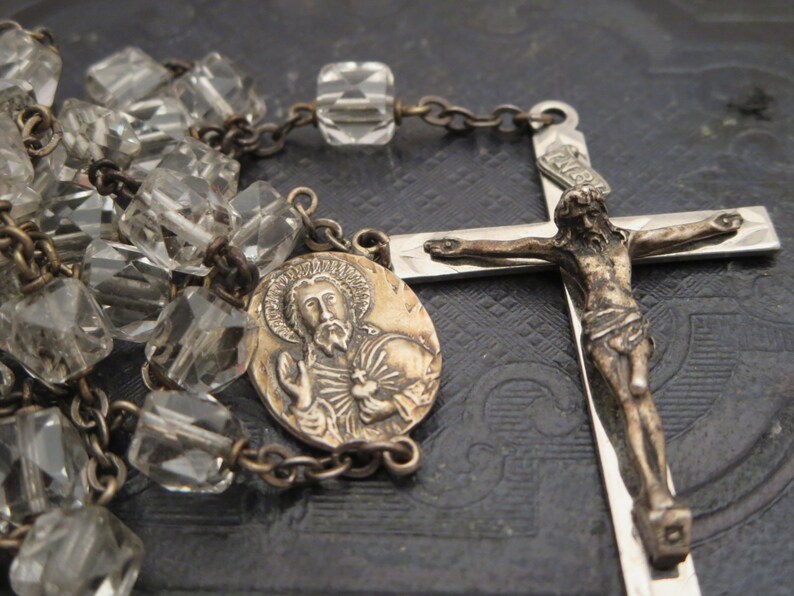 May include: A silver rosary with a clear glass bead chain, a silver crucifix with a figure of Jesus Christ, and a silver medallion with a depiction of a saint.
