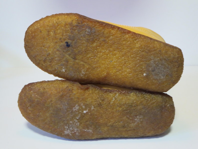 May include: Close-up of the soles of two yellow suede shoes stacked. The soles are textured, light brown rubber with white marks. The shoes appear new. The image shows the underside of the shoes.