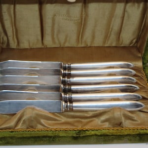 May include: A set of six silver butter knives in a green velvet lined box. The knives are arranged in a row and have a polished finish.